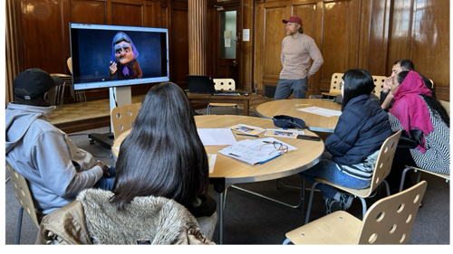 A group of six people are gathered around round tables in a wood-panelled room. They are all looking to a large TV screen that is showing a scene from an animated film. 