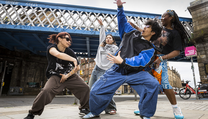 Four street dancers perform expressive, energetic moves beneath a blue iron bridge in an urban area. They wear casual, loose-fitting clothes and sneakers, striking dynamic poses that convey rhythm and creativity. Behind them, stone buildings and a pink sign reading “People Make Glasgow” highlight the lively city backdrop.