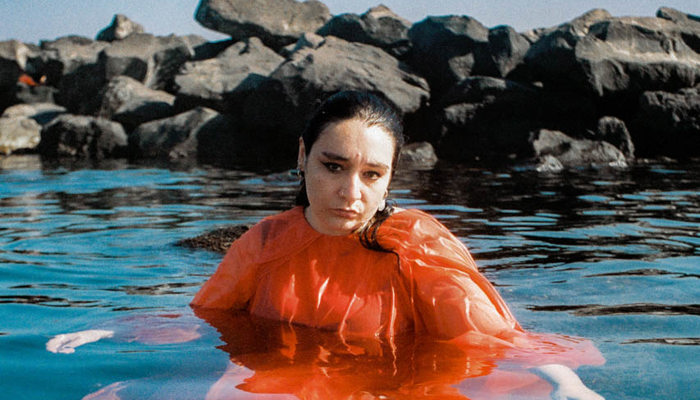 A person stood in water wearing a orange stop with wet, slicked back hair. 