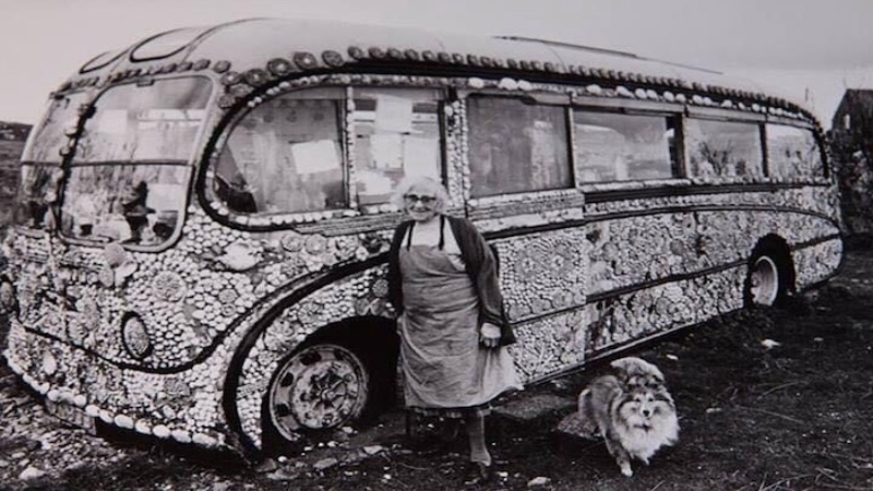Black and white image of Flora with Shell the dog. They are standing in front of her bus that has been completely decorated in shells.
