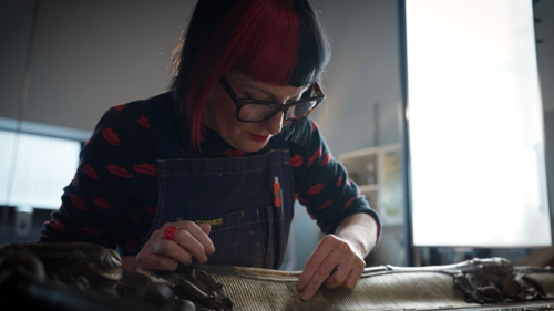 In a workshop, lit by a special lamp, frame conservator with black rimmed glasses and red and black bobbed hair, wearing a denim work apron over a dark blue jumper with red lips, has her head down carefully applying gold leaf to a frame.