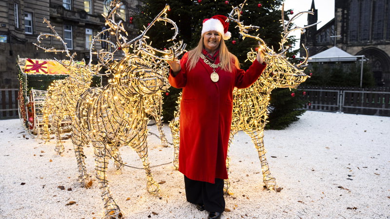 A person in a red coat and Santa hat standing among illuminated wire-frame reindeer decorations in a snowy outdoor setting, with a large Christmas tree and historic buildings in the background