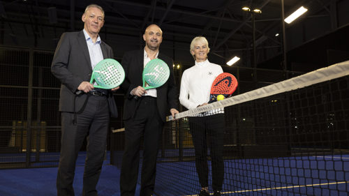 Two men and a woman stand either side of the net on a padel holding padel racquets, the woman is also holding a padel ball