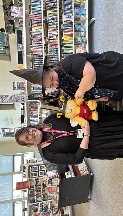 Two people dressed in Halloween costumes standing in front of a book display in a library. One is a bat, the other a witch. They are holding a yellow stuffed toy between them.