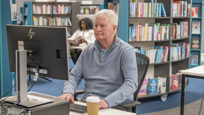 A person sitting typing at a computer in a library with with bookshelves behind them.