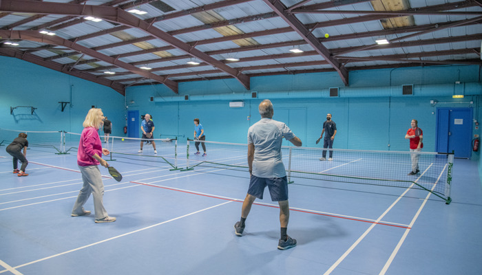 A wide view of two sets of people playing pickleball doubles across two indoor courts which are a blue hard surface.