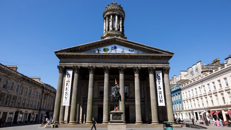 The Gallery of Modern Art in Glasgow, a neoclassical building with tall columns and a central dome. Two large banners hang on the façade—one reading “Banksy” with a traffic cone replacing a letter, and the other reading “Cut & Run.” In front of the building stands the Duke of Wellington equestrian statue, with orange traffic cones placed on the rider’s head and the horse’s raised leg.