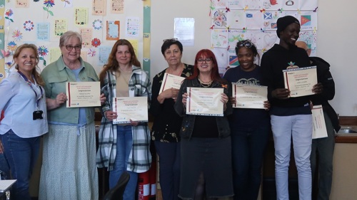 A group of seven adult learners are gathered for a photo in front of a learning display in a community centre. They are all holding certificates