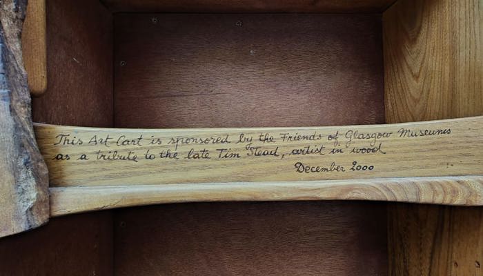 A wooden shelf with a written text: "This Art Cart is sponsored by the Friends of Glasgow Museums as a tribute to the late Tim Stead, artist in wood. December 2000"