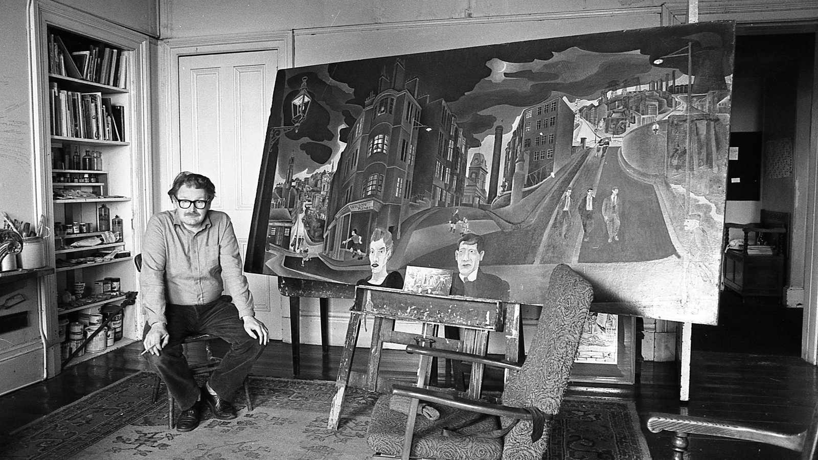 A black‑and‑white photograph of an artist seated beside a large painting on an easel. The artist, wearing glasses and a button‑up shirt, sits in a chair holding a cigarette. Shelves filled with paint cans and brushes line the left side of the room. The painting depicts a distorted urban landscape with looming buildings, dark clouds, streetlamps, small human figures, and two large faces at the bottom. The studio has a patterned rug, wooden floors and some furniture, creating an intimate, lived‑in workspace.