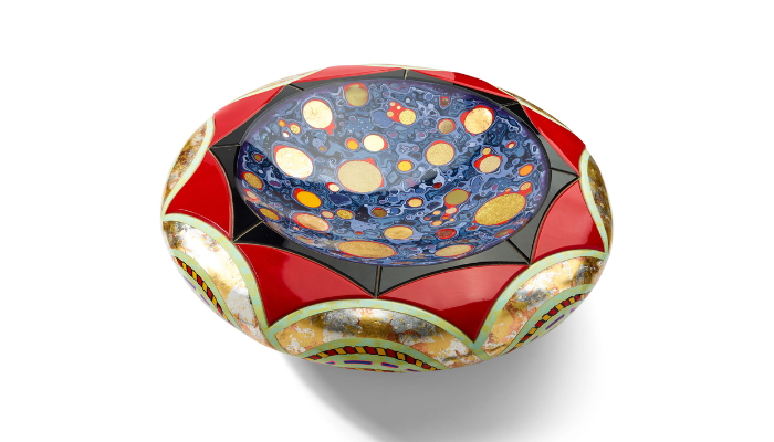 Decorative round bowl with a vibrant red exterior and an intricately patterned interior. The inside features a blue marbled surface filled with gold and cream circular shapes of varying sizes, while black geometric sections separate the colourful panels. Metallic gold, silver, and copper detailing around the rim adds a reflective, ornate finish. The bowl is photographed against a plain light background.