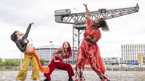Dancers wearing multicoloured outfits strike poses in front of a large crane and music venue in the background