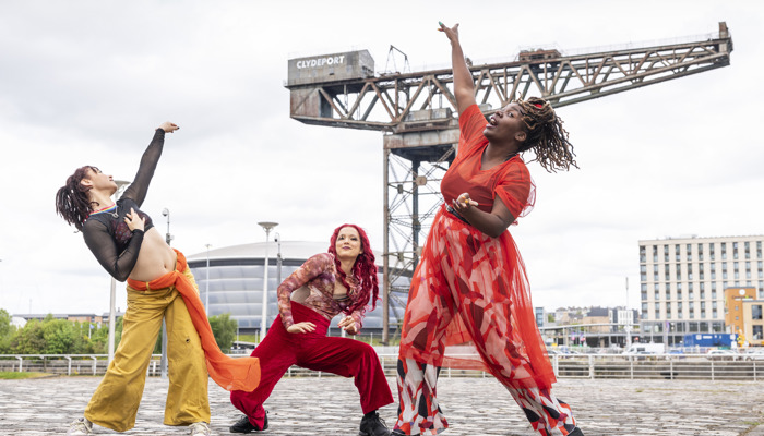 Dancers wearing multicoloured outfits strike poses in front of a large crane and music venue in the background